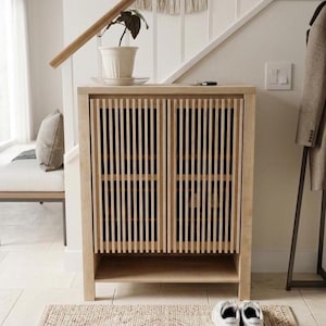 May include: A light-colored wooden cabinet with vertical slat doors and a lower shelf. A small plant in a white pot sits on top. White sneakers rest on a woven rug in front of the cabinet. A coat rack and stairs are in the background.