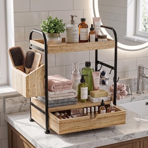 May include: A two-tiered wooden bathroom organizer with black metal supports. The top shelf holds a potted plant and various skincare products. The lower shelf features a drawer with makeup and a compartment for towels and toiletries. Brushes are stored in a wooden container.