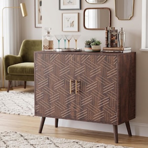 May include: A dark brown wooden cabinet with two doors featuring a geometric pattern of thin gold lines. The cabinet stands on four tapered legs and has two vertical gold handles. The top surface holds a glass decanter, four cocktail glasses, a small potted succulent, and three books.