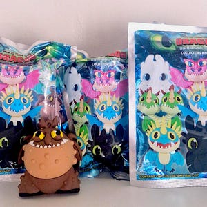Peut inclure: Plusieurs clips de sac à collectionner avec des personnages de "Dragons". Les clips sont dans des sacs scellés avec un design bleu et vert. Un clip de personnage marron est en dehors du sac. Les sacs ont le texte "DRAGON" et "COLLECTORS BAG CLIP."