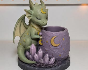 May include: A sage green dragon figurine with closed eyes, featuring a gold crescent moon on its forehead and a matching necklace. It stands beside a textured purple cylindrical container adorned with a gold crescent moon and purple crystals.