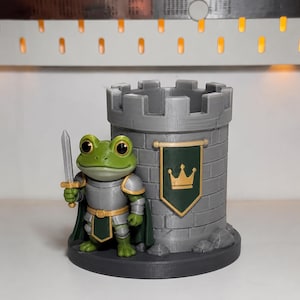 May include: A gray cylindrical pencil holder shaped like a castle tower with a crenelated top. A green frog knight in silver armor stands in front, holding a silver sword. A green shield with a gold crown emblem is attached to the tower wall.