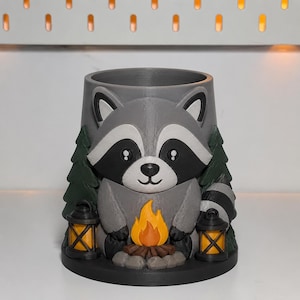 May include: A gray cylindrical container featuring a sculpted raccoon sitting in front of a campfire. The design includes dark green pine trees on either side and two small black lanterns with orange flames at the base of the raccoon figure.