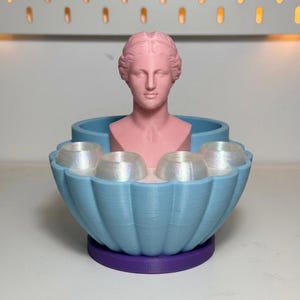 Puede incluir: Pieza decorativa impresa en 3D con un busto rosa de una figura clásica sobre un recipiente en forma de concha azul claro. El recipiente contiene cuatro objetos iridiscentes en forma de cúpula. La base es morada.