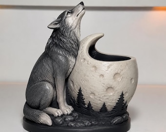May include: A gray and white wolf figurine sitting and howling toward the sky, positioned next to a textured, cratered moon-shaped container. The base features a black forest silhouette with pine trees and rocks on a circular black platform.