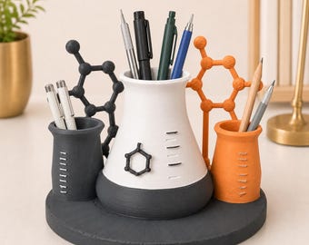 May include: A desk organizer featuring a central white and dark gray conical flask-shaped pen holder, flanked by a smaller dark gray and orange cylindrical holder. The set includes black and orange molecular structures and holds various pens and pencils.