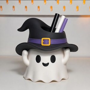 May include: A white ghost-shaped desk organizer featuring a black witch hat with a purple band and a yellow buckle. The ghost has two black oval eyes and a small black smiling mouth. Three pens in black, white, and purple are stored inside.