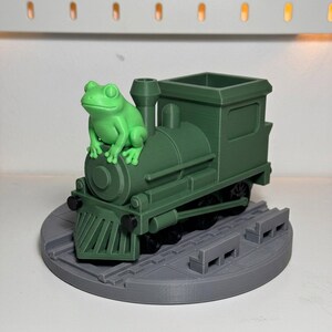 May include: A green 3D-printed toy train with a frog figurine perched on top. The train is dark green and sits on a gray circular base with train tracks. The frog is bright green.