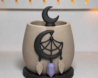 May include: A beige cylindrical planter with a black crescent moon topper and a black crescent moon dreamcatcher emblem on the front. The emblem features two beige feathers and a central light purple crystal pendant, all resting on a black base.