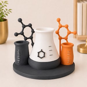 May include: A desk organizer featuring a central white and dark gray conical flask, flanked by a dark gray cylindrical cup and an orange cylindrical cup. Each cup has measurement markings. A black and an orange molecular structure model are attached.