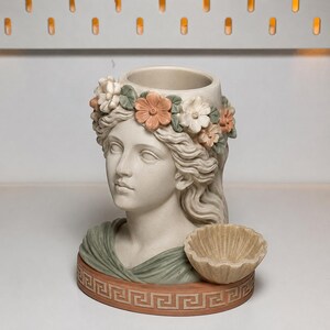 May include: A beige ceramic bust planter featuring a sculpted face adorned with a crown of pastel pink, white, and green flowers. The base is decorated with a terracotta-colored Greek key pattern and includes a small, attached shell-shaped dish.