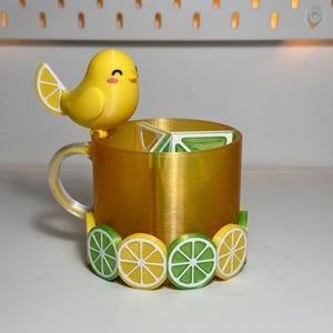 May include: A yellow 3D-printed mug with a clear handle and a decorative bird perched on the rim. The mug is adorned with lemon and lime slice accents around the base. The bird has a lemon slice wing.