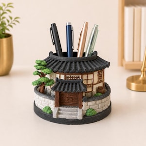 May include: A circular desk organizer shaped like a traditional East Asian building with a dark gray tiled roof, white walls, and a brown wooden gate. The structure features a small green tree and stone-like walls, holding four pens.