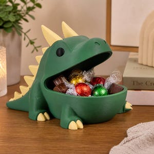 May include: A dark green dinosaur-shaped bowl with a wide-open mouth containing several wrapped candies. The creature features light yellow spikes along its back and tail, small yellow claws on its feet, and two solid black circular eyes.