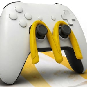 以下が含まれることがあります： 黒いジョイスティックとボタンが付いた白いゲームコントローラー。黄色の「M」のデザインが特徴です。 コントローラーは、ファストフードをテーマにした白と黄色のストライプの紙の上に置かれています。