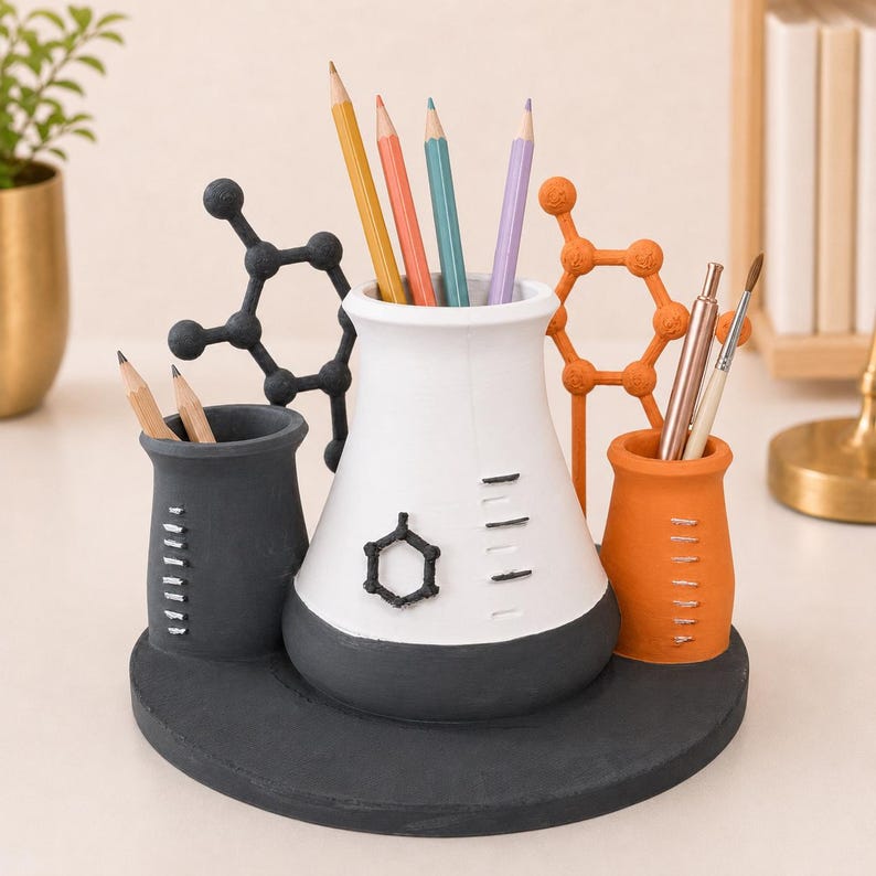 May include: A desk organizer shaped like laboratory beakers and flasks on a circular base. The central white flask holds pencils, flanked by a smaller black beaker and an orange beaker. Decorative black and orange molecular structures are attached.