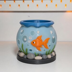 May include: A light blue, bowl-shaped container featuring a dark blue rim, a bright orange goldfish relief, and cut-out circular bubble shapes. The base is decorated with sculpted green seaweed and grey stones, all set upon a dark grey circular stand.