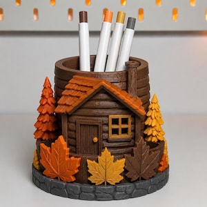 May include: A cylindrical brown desk organizer shaped like a log cabin with a chimney, surrounded by sculpted orange, yellow, and brown maple leaves and evergreen trees. Four markers with white caps are stored inside the central compartment.