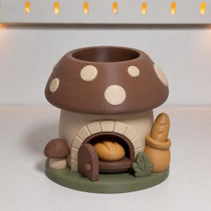 Op de afbeelding: Een bruine paddestoelvormige houder met crèmekleurige stippen op de hoed. De basis heeft een boogvormige deuropening met een open deur, een brood binnenin, een kleine paddestoel links, een groen blad en een zak met een stokbrood.