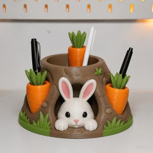 May include: A brown tree stump desk organizer featuring a white rabbit peeking out from a hollow opening. The organizer includes three orange carrot-shaped pen holders with green tops, each containing a pen, and green grass accents at the base.