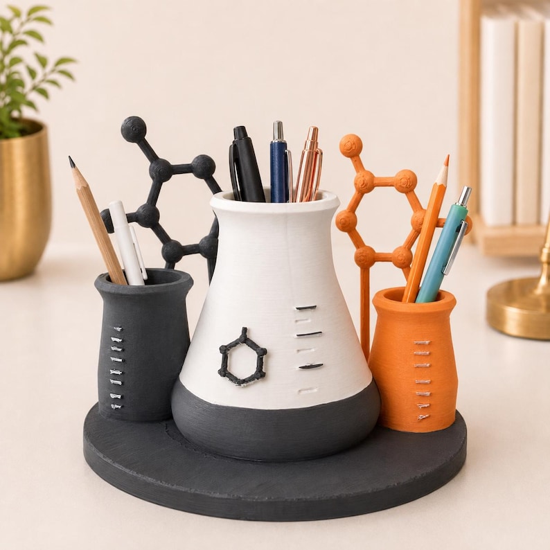 May include: A desk organizer featuring a central white and dark gray conical flask-shaped container, flanked by a smaller dark gray cylinder and an orange cylinder. Each container has measurement markings. Decorative black and orange molecular structures stand behind the containers, all mounted on a circular dark gray base.