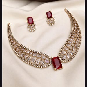 Può includere: Un set di collana e orecchini color oro con una pietra centrale rettangolare rosso scuro, affiancata da due ali curve riempite di cristalli trasparenti a goccia e piccole pietre rotonde. Gli orecchini abbinati presentano una singola pietra rossa.