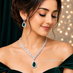 May include: A silver-toned necklace and matching drop earring set featuring pear-shaped emerald green stones surrounded by a halo of clear white crystals. The necklace chain consists of circular crystal links leading to a central teardrop pendant.