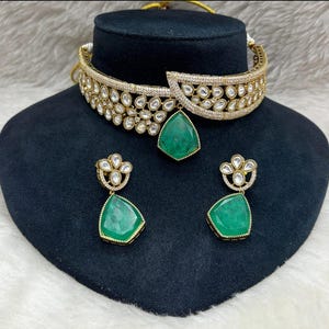 Könnte beinhalten: Ein goldfarbenes Halsketten- und Ohrring-Set mit klaren und smaragdgrünen Akzenten. Die Halskette im Choker-Stil und die Ohrringe haben ein Blumenmuster mit klaren Steinen und einen großen, tropfenförmigen smaragdgrünen Anhänger.