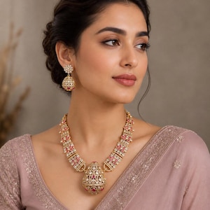 May include: A gold-toned necklace and matching earrings set. The necklace features a pendant with red and clear stones. The earrings have a similar design. The jewelry is worn by a person wearing a pink garment.