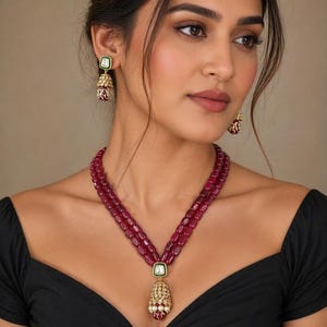 Può includere: Un set di collana e orecchini con perline rosso rubino. La collana presenta un pendente con una pietra quadrata bianca, filigrana dorata e un rubino a forma di goccia. Gli orecchini hanno un design simile con una pietra quadrata verde e dettagli di rubino.