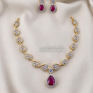 Può includere: Un set di collana e orecchini color oro con pietre rosse a goccia circondate da piccoli cristalli bianchi. La catena della collana è composta da maglie a forma di ala con cristalli bianchi e un ciondolo centrale a goccia.