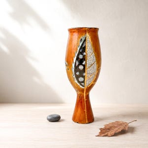 May include: A tall, amber-colored wooden vase with a flared rim and a narrow base. The exterior features a decorative inlay shaped like a leaf, divided into two sections: one with dark, reflective mosaic pieces and one with a white, crackled texture.