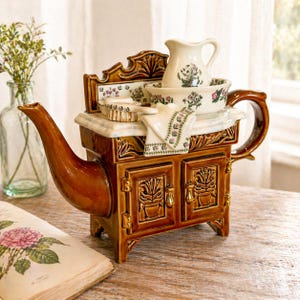 Könnte beinhalten: Eine braune Keramik-Teekanne in Form eines Vintage-Ofens mit Ausguss und Griff. Sie hat eine dekorative Rückwand und Schranktüren. Eine kleine Kanne, eine Schüssel und andere Gegenstände mit floralen Mustern liegen obenauf. Ein dekoratives Handtuch ist ebenfalls vorhanden.