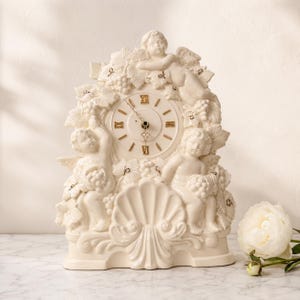 May include: An off-white ceramic mantel clock featuring three sculpted cherubs, grape clusters, and vine leaves. The clock face has gold Roman numerals and thin black hands. A large shell motif is centered at the base of the clock structure.