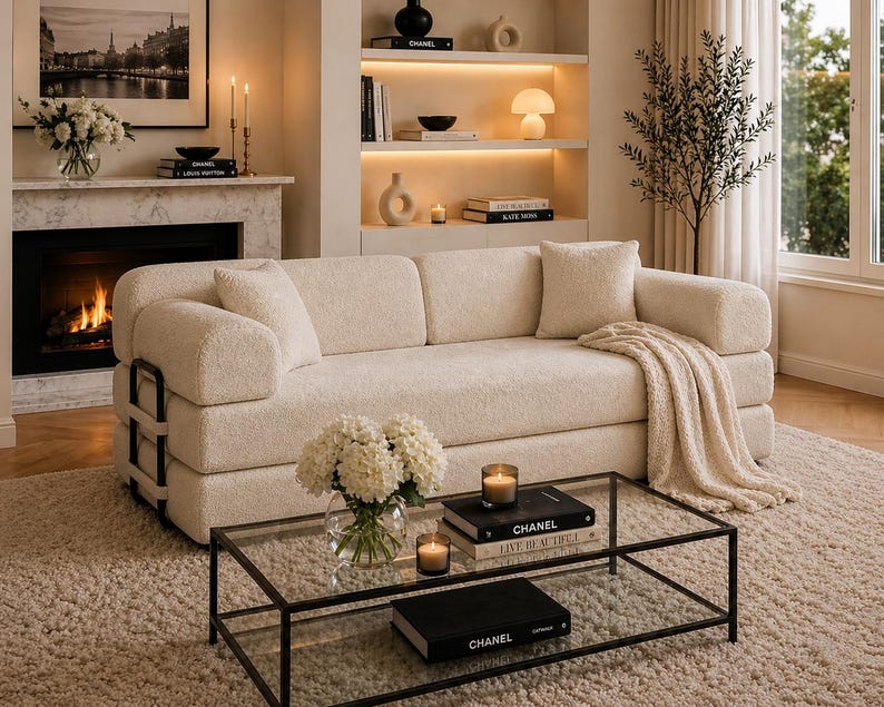 May include: A cozy living room scene featuring a cream-colored sofa with black metal accents, a glass coffee table, and a fireplace. The room is decorated with candles, flowers, and books, creating a warm and inviting atmosphere.