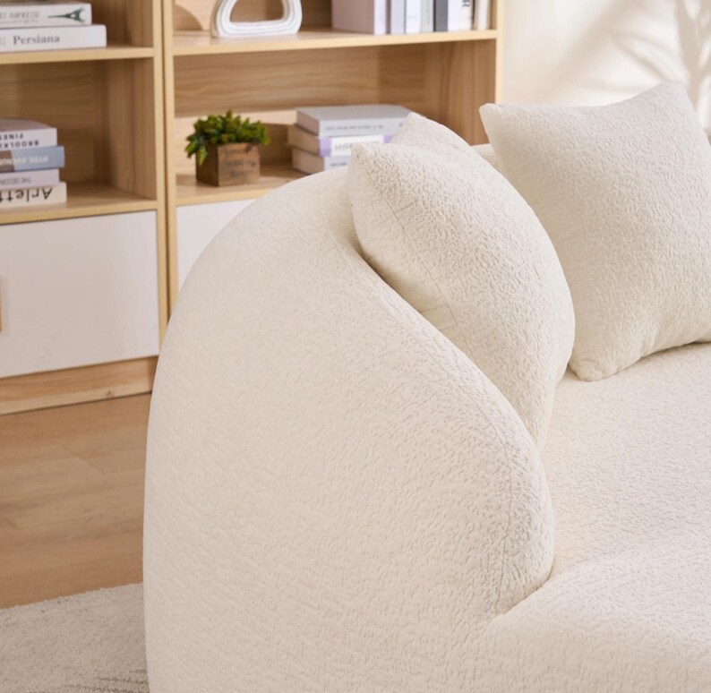 May include: Cream-coloured, curved sofa with a textured, plush fabric. Two matching throw pillows are visible. The sofa is positioned in front of a light-coloured wooden bookshelf.