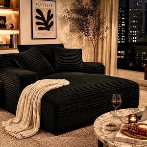 May include: A black corduroy sectional sofa with several matching pillows. A cream-colored knitted throw blanket is draped over the sofa. A small marble side table with a lamp and a candle sits next to the sofa. A coffee table with a laptop, wine, and snacks is in front of the sofa.