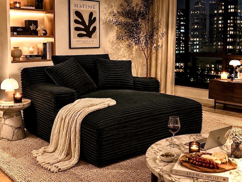 May include: A black corduroy sectional sofa with several matching pillows. A cream-colored knitted throw blanket is draped over the sofa. A small marble side table with a lamp and a candle sits next to the sofa. A coffee table with a laptop, wine, and snacks is in front of the sofa.
