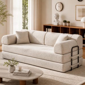May include: A cream-colored, modern sofa with rounded armrests and a matching square pillow. The sofa has a textured fabric and a black metal frame on the side. The room features a neutral color palette with a rug, coffee table, and wooden shelving.