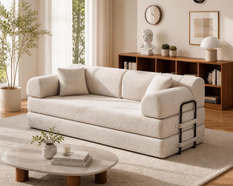 May include: A cream-colored, modern sofa with rounded armrests and a matching square pillow. The sofa has a textured fabric and a black metal frame on the side. The room features a neutral color palette with a rug, coffee table, and wooden shelving.