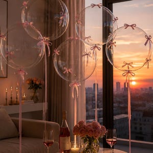 May include: Transparent balloons with pink ribbons and bows float near a window with a sunset view. A table is set with wine, flowers, candles, and a bottle of rosé, creating a romantic scene.