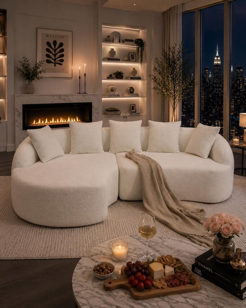 May include: An elegant living room with a large, cream-coloured sectional sofa and cushions. A marble coffee table displays a charcuterie board, wine, and a candle. A fireplace and city skyline view create a sophisticated, inviting atmosphere.