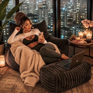 May include: A cozy indoor scene featuring a large, dark gray beanbag chair and matching ottoman. A person is relaxing, wearing a white crop top and dark leggings, using a smartphone. A laptop sits on the ottoman. A soft, beige blanket and a small table with snacks and candles complete the scene.