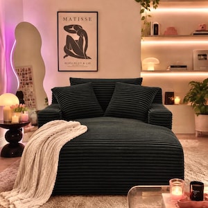 May include: A dark green corduroy sofa with two matching pillows and a cream-colored throw blanket. The sofa is in a living room with a rug, a mirror, and a framed Matisse print. Candles and decorative items are on a side table and shelves.