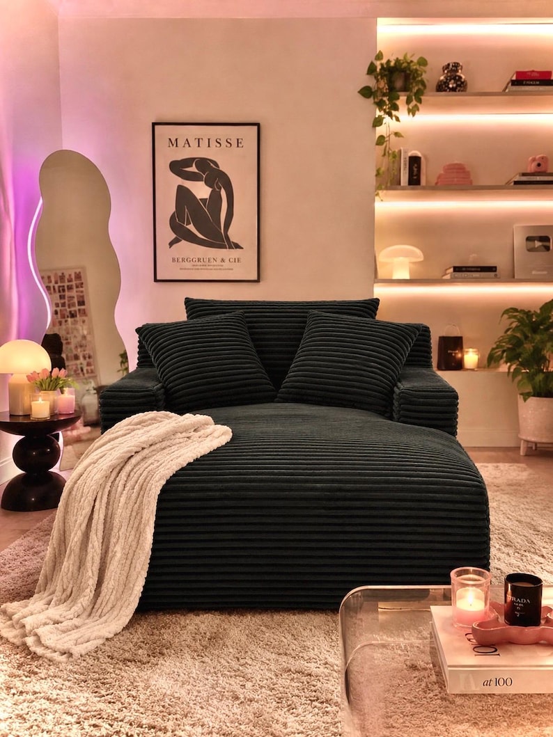 May include: A dark green corduroy sofa with two matching pillows and a cream-colored throw blanket. The sofa is in a living room with a rug, a mirror, and a framed Matisse print. Candles and decorative items are on a side table and shelves.