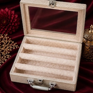May include: A rectangular jewelry display case with a shimmering champagne-colored exterior and a clear glass lid. The interior features three soft, beige velvet-lined ring rolls for organization, secured by a silver-toned metal latch and handle.