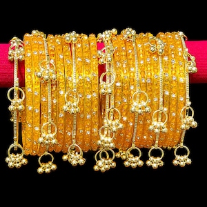 Könnte beinhalten: Ein Set aus gelben Armreifen mit strukturierter Oberfläche und eingebetteten klaren Kristallen. Jeder Armreif ist mit goldfarbenen Metallanhängern verziert, die aus kleinen Ringen und Trauben winziger Kugeln bestehen.