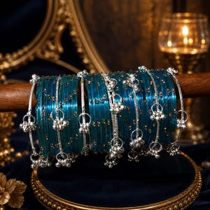 Può includere: Un set di braccialetti rigidi color blu ottanio traslucido con piccole macchie dorate incastonate nel materiale. I braccialetti sono decorati con catenelle in metallo argentato che pendono verticalmente, terminando con piccoli grappoli di campanellini argentati.