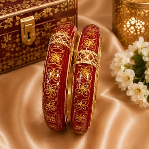 May include: Two circular bangles featuring a deep red glossy finish with intricate gold-toned floral patterns and a central lattice-style gold band. The bangles have a smooth, rounded profile and a reflective surface, showcasing detailed metalwork.