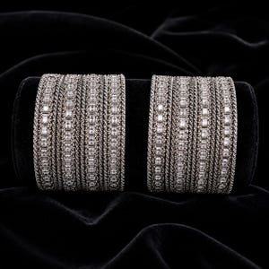 Könnte beinhalten: Zwei breite Armreifen mit dunklem Metallic-Finish, bestehend aus mehreren Reihen texturierter, seilartiger Bänder, die sich mit Reihen rechteckiger, klarer Steine abwechseln. Das Design besteht aus mehreren gestapelten Armreifen.